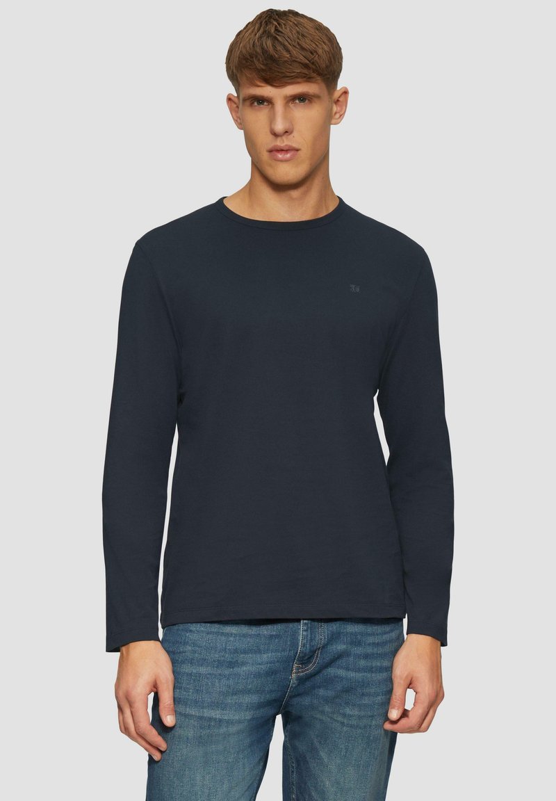 Marineblaues Langarm-T-Shirt aus Baumwolle, mit rundem Ausschnitt, geradem Saum und einem kleinen Logodetail auf der Brust. Trägt dazu eine blaue Jeans.