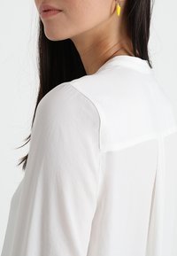 Witte blouse met een gladde textuur, met een ronde halslijn en een contrasterend achterkantdetail. Een model draagt gele oorbellen.
