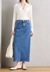 Camisa blanca de manga larga, falda larga de mezclilla azul de cintura alta, zapatos planos crema con pequeños acentos de hardware y un bolso color beige. Fondo de piso de madera.