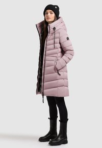 Kvinde iført en lang mauve pufferjakke med hætte, sort beanie, sorte leggings og sorte kraftige støvler, stående med hænderne i lommene på jakken.