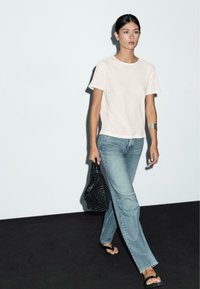 Hvid bomulds T-shirt, lyseblå jeans, sort vævet totebag og sorte flip-flops; model der går mod en neutral baggrund.