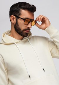 Crème hoodie met witte trekkoorden, voorzien van een voorzak. Zwarte zonnebril met oranje lenzen. Halflang golvend haar en een baard.
