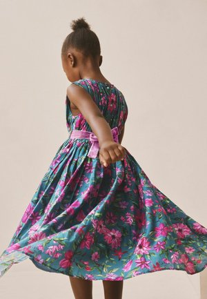 Junges Mädchen dreht sich, trägt ein ärmelloses türkises Kleid mit pinkem Blumenmuster und einem pinken Samtschleifengürtel, vor einem einfarbigen beigen Hintergrund.