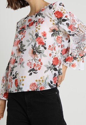 Femme portant un chemisier blanc avec un imprimé floral rouge et rose et des détails en dentelle sur les manches évasées, associé à un pantalon noir.