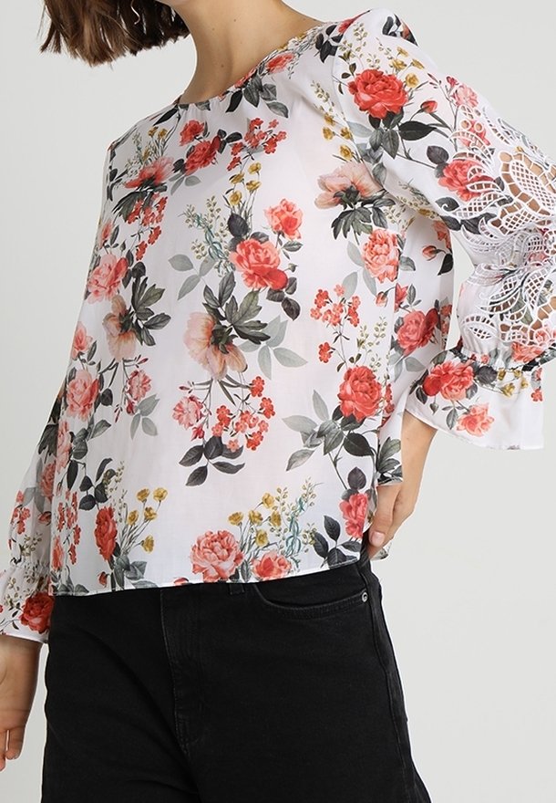 Femme portant un chemisier blanc avec un imprimé floral rouge et rose et des détails en dentelle sur les manches évasées, associé à un pantalon noir.