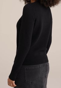 Svart texturerad tröja med långa ärmar och ribbad nederkant, bärs med svarta jeans. Har en figurnära design och en subtilt mönster.