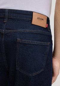 Dunkelblaue Denim-Jeans mit einem sichtbaren Lederpatch mit der Aufschrift "JOOP! JEANS" und kontrastierenden orangen Nähten. Standard-Design der Gesäßtasche.