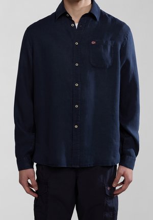Mand iført en mørkeblå skjorte med knapper og et lille flagemblem på brystlommen samt matchende mørkeblå cargobukser.