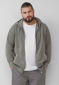 Grijze rits-hoodie met een trekkoordkap, frontzakken en contrasterende stiksels, gedragen over een wit T-shirt en grijze broek.