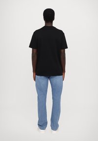 T-shirt noir à manches courtes avec col rond, porté avec un jean bleu clair et des baskets blanches, vu de dos sur un fond uni.