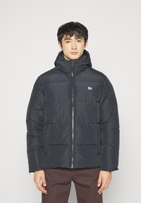 Veste matelassée bleu foncé avec capuche, présentant des sections quiltées, fermeture à glissière et logo brodé sur la poitrine gauche.