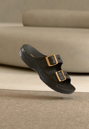 Czarny sandał typu slip-on z dwoma regulowanymi złotymi sprzączkami, teksturowaną wkładką i grubą podeszwą, unoszący się nad beżowym dywanem w neutralnym pomieszczeniu.