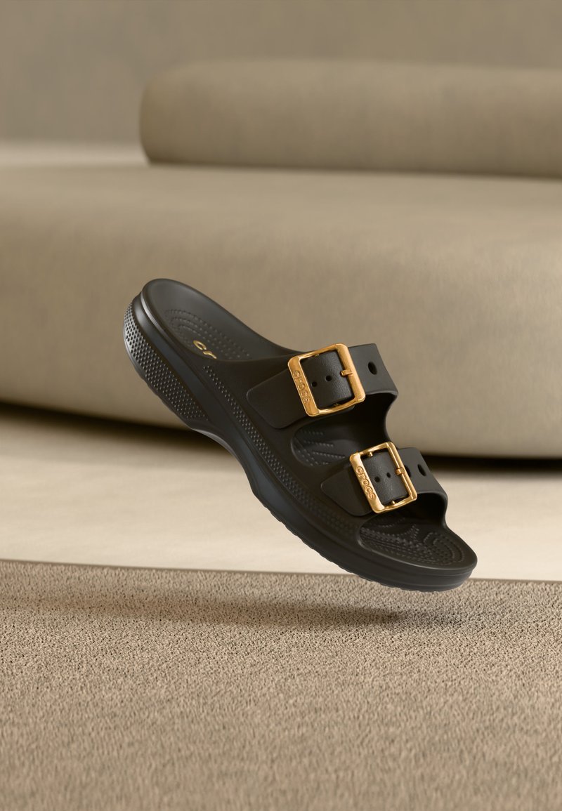 Sandale noire à enfiler avec deux boucles dorées réglables, semelle intérieure texturée et semelle épaisse, flottant au-dessus d'un tapis beige dans une pièce neutre.