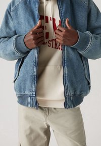 Ljusblå denim bomberjacka med ribbade muddärmar och dragkedja, lager över en krämfärgad hoodie med röd text. Krämfärgade byxor fulländar outfiten.