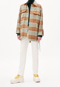 Person klädd i vita byxor, en överdimensionerad beige och grön rutig jacka över en mörkgrön topp, samt vita sneakers med gula snören mot en vit bakgrund.