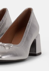 Scarpe con tacco argentato con finitura metallica texturizzata, punta arrotondata e tacco a blocco con base nera liscia. Accentuate da un dettaglio a fiocco.