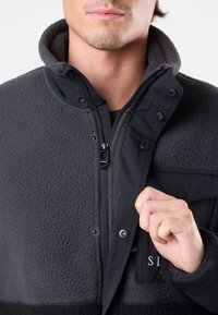 Giacca in fleece nera e grigia con bottoni a pressione e chiusura con cerniera. Presenta una texture morbida e una tasca sul petto con dettaglio stampato.