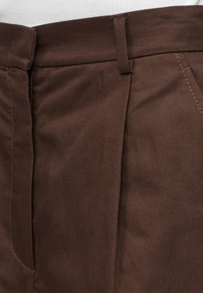 Κοντινή λήψη της μέσης σκούρων καφέ παντελονιών με θηλιές ζώνης, πιέτα μπροστά και ραφή πλαϊνής τσέπης σε λευκό υφασμάτινο φόντο.