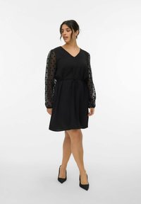 Zwarte jurk met een V-hals en een tailleriem, met doorzichtige lange mouwen met stippen en een soepele rok. Draagt het met zwarte puntige hakken.