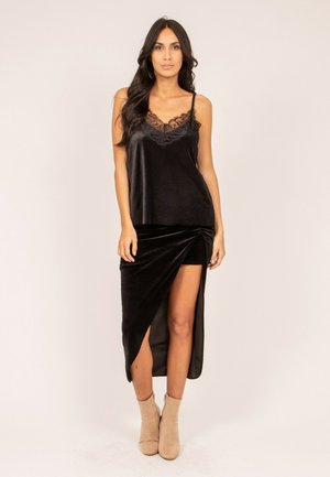 Femme aux longs cheveux noirs portant un caraco noir orné de dentelle et une jupe asymétrique en velours noir avec des bottines beiges, debout devant un fond uni.