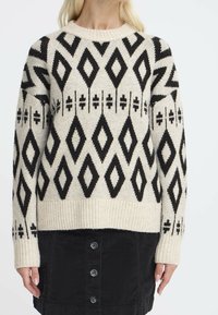 Donna che indossa un maglione beige con motivi geometrici a diamante neri e una gonna nera con bottoni su uno sfondo bianco uniforme.