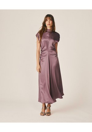 Mujer con vestido midi morado brillante sin mangas con fruncidos en los laterales y sandalias plateadas de tacón alto con tiras, de pie sobre un fondo liso.