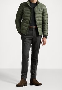 Mężczyzna w oliwkowej pikowanej kurtce noszący pod nią szary sweter, szare eleganckie spodnie, brązowy pasek oraz brązowe zamszowe buty, stojący z rękami częściowo w kieszeniach.