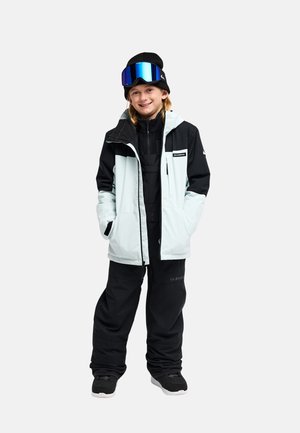 Jonge persoon in een zwart-witte skijas, zwarte broek, zwarte muts en blauwe skibril glimlacht met handen in de jaszakken.