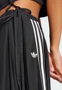 Close-up of a person wearing black Adidas pants with white stripes and logo, showing waist and hand resting by the side.