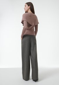 Rippierter, mauvefarbener Pullover mit einer geschichteten Rüsche auf der Rückseite, kombiniert mit hoch taillierten, weit geschnittenen grauen Hosen, die seitliche Taschen haben.