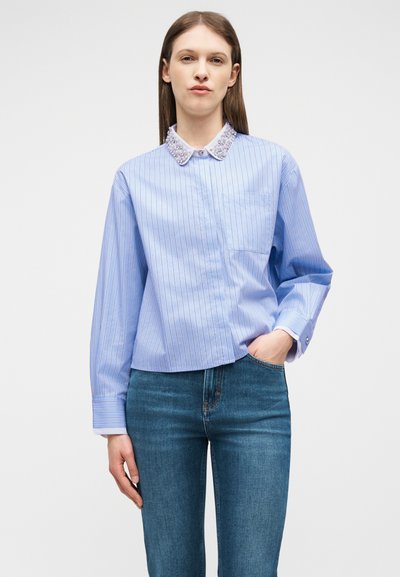 Νεαρή γυναίκα που φοράει μπλε ριγέ πουκάμισο με διακοσμημένο γιακά και μπλε τζιν παντελόνι, στέκεται με το ένα χέρι στην τσέπη, σε απλό φόντο.