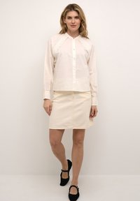 Lysecremfarvet skjorte med knapper og en bølget blondedetalje, parret med en cremefarvet denimnederdel. Sorte flade sko fuldender outfittet.