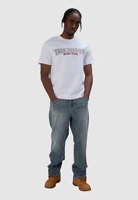 Witte katoenen T-shirt met het logo "TRUE RELIGION BRAND JEANS" in grijs en rood. Blauwe spijkerbroek en bruine laarzen maken de outfit compleet.