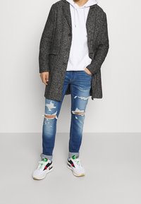 Grauer strukturierter langer Mantel über einem weißen Hoodie, kombiniert mit ausgewaschenen blauen Jeans und weißen Sneakers mit schwarzen und roten Akzenten.