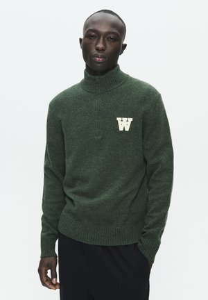 Dark green knitted sweater with a high collar and quarter-zip. Features a cream-colored letter "W" on the left chest. Textured fabric.