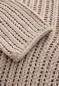 Strikket beige stof med lodrette åbne masker og skinnende accenter, med en foldet kant og et blødt, struktureret udseende.