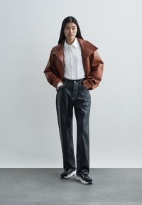 Casaco oversized de couro castanho, camisa branca, calças pretas de couro sintético e ténis desportivos pretos com detalhes brancos, em pé contra um fundo claro.