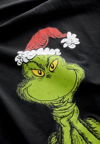 Melnā T-krekla attēlā redzams zaļš Grinčas tēls ar dzeltenām acīm, valkājot sarkanu un baltu Ziemassvētku cepuri, uz gludas auduma fona.