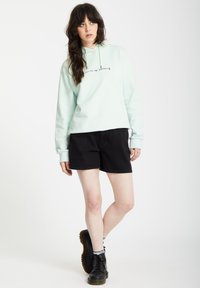 Felpa con cappuccio azzurra chiara con stampa grafica, abbinata a pantaloni corti neri e stivali neri stringati con suola spessa.
