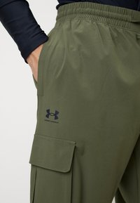 Pantalon cargo vert olive avec une ceinture élastique, des poches latérales et un logo noir Under Armour sur la cuisse. Tissu lisse et léger.