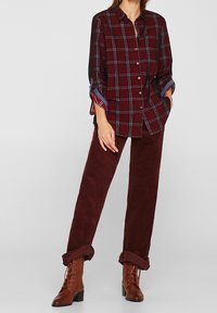 Camicia a quadri bordeaux con dettagli blu, abbinata a pantaloni in velluto a coste bordeaux e stivali alla caviglia marroni con lacci. Tutti gli articoli sono casual.