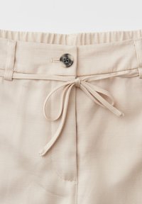 Beige broek tailleband met elastische achterkant, enkele schildpaddenknop, lussen voor een riem en een geknoopte stoffen trekkoord aan de voorkant.