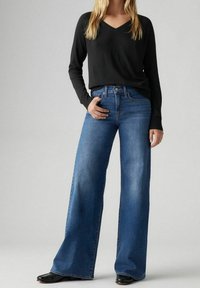 Szerokie niebieskie dżinsy z wysokim stanem, z pięcioma kieszeniami i zapięciem na guzik, zestawione z czarną, długorękawową bluzką z dekoltem w serek.