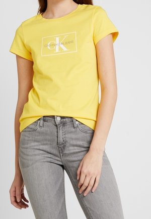Personne portant un t-shirt jaune Calvin Klein Jeans et un jean gris clair, une main posée sur la cuisse et l'autre détendue.