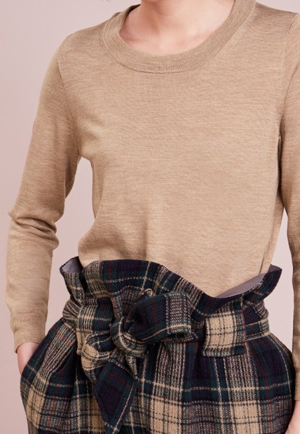 Femme portant un pull beige à manches longues rentré dans une jupe taille haute à carreaux bleu marine et beige avec une grande ceinture nouée en forme de nœud.