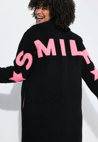 Giacca di pile nera con grande scritta rosa che dice "SMILE" sulle maniche. Texture morbida, maniche lunghe e vestibilità comoda.
