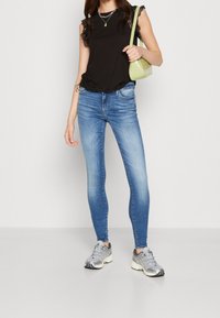 Top negro sin mangas con encaje, vaqueros ajustados azules y zapatillas deportivas grises. Sosteniendo un bolso verde pequeño.