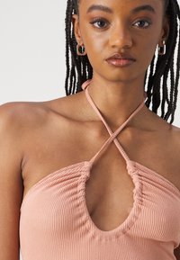 Haut halter en rose clair côtelé avec un décolleté en V profond et des détails froncés, présentant des straps croisées au niveau du cou et une texture lisse.