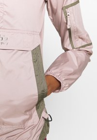 Main d'une femme dans la poche d'une veste rose clair et vert olive avec un poignet élastique et des détails de poche zippée sur le bras.