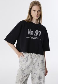 T-shirt noir raccourci à manches courtes ornée de gros caractères blancs "N° 97" et de graphismes détaillés, associée à un pantalon gris clair recouvert de motifs variés.